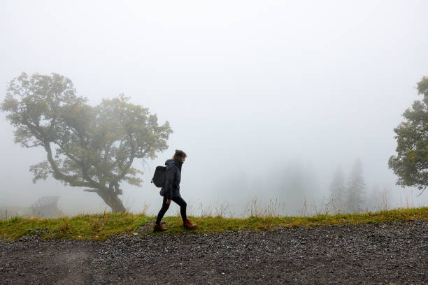 Lonely female hiker going through thick mist while carrying a backpack and a jacket in the Austrian Alps