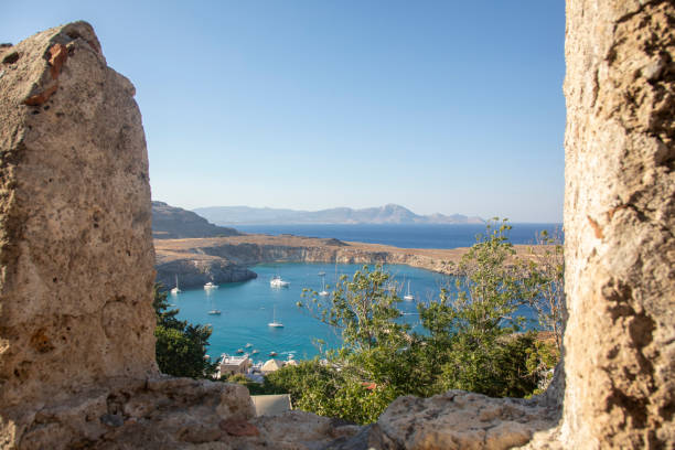 Elevated shot looking through a stone wall of a bay of water with rocky outcrop on Rhode Island, Greece. There are small sailing boats bobbing in the bay and in the far distance a hilly horizon is visible, beyond the deep blue sea.