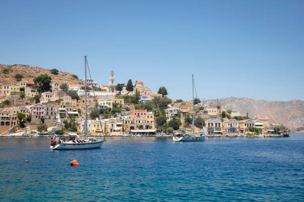 Medium shot looking across a bay of water to a traditional town set on a hill on the island of Symi, Rhodes, Greece. It is a bright sunny day and there are sailing boats floating in the bay. The traditional buildings are a variety of different pastel colours.