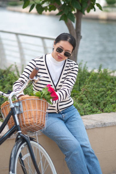 A stylish woman in a striped sweater holds a bouquet of pink roses arranging them on a bike basket while seated by a bicycle, creating a beautiful and serene outdoor scene. The vibrant colors enhance the mood.