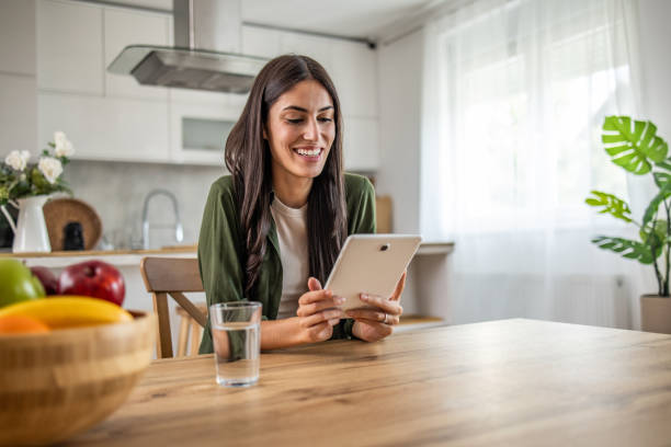istock Smiling young woman using digital tablet while sitting at table in dining room 2189560485