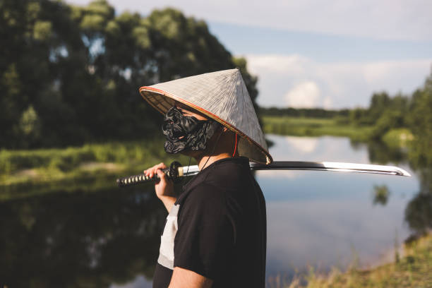 istock A man wearing a samurai mask, conical hat and black shirt holds a katana on his shoulder while standing by a river, representing a warrior or historical reenactor. Samurai warrior on the river bank. 2189563783