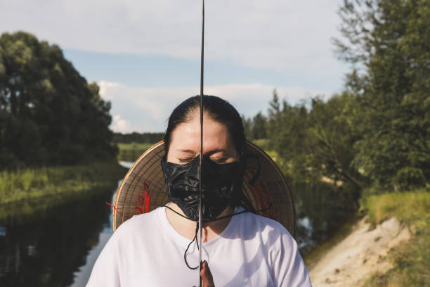 istock Female cosplayer with closed eyes wearing black samurai mask, conical straw hat and holding katana in front of face in nature near river. Samurai with katana in threatening pose. 2189566340