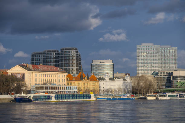 Riverside scene in Bratislava featuring historic and modern architecture, docked riverboats, and reflective water under a cloudy sky.