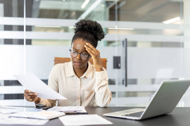 istock Young professional woman in modern office reviewing financial documents with thoughtful expression, surrounded by laptop and paperwork, illustrating business challenges and productivity 2189580904