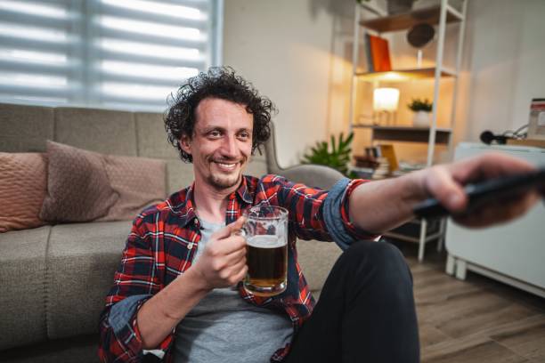 istock Young man drinking beer and watching tv at home 2189581505