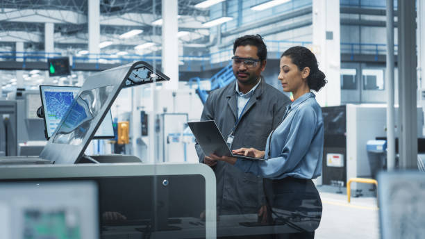 istock Indian Male Industrial Engineer And Hispanic Female Manager Using Laptop And Talking At Electronics Factory. Man Using Soldering Jet Printer, Explaining Process Behind Production Of New Motherboards. 2189585694