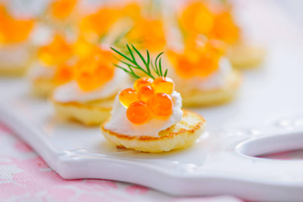 Close-up of mini blinis topped with sour cream, salmon roe, and fresh dill on a decorative white platter