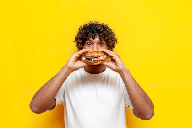 hungry indian guy eats a delicious burger on a yellow isolated background and is surprised