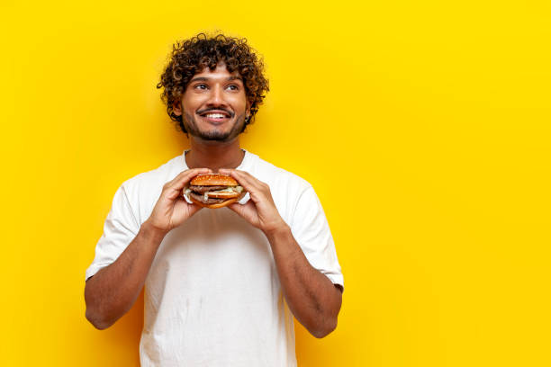 istock pensive indian guy eating a delicious burger on a yellow isolated background and thinking 2189588590