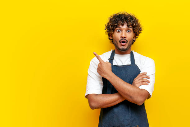 istock young surprised guy indian waiter in apron pointing at copy space on yellow isolated background 2189588631