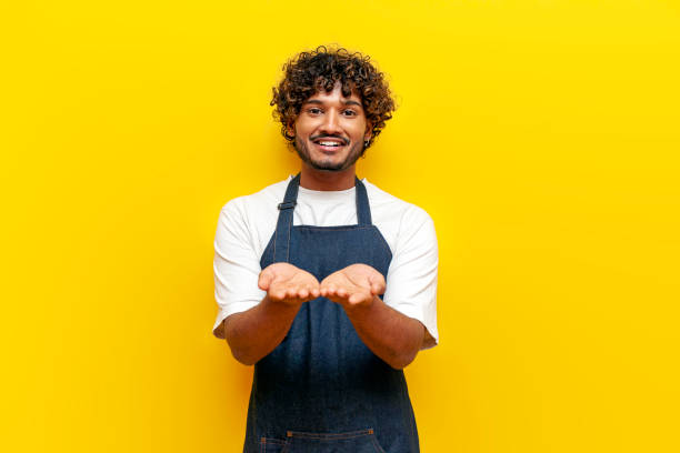 young surprised guy Indian waiter in an apron begs on a yellow isolated background, male Hindu barista in uniform holds empty hands and gives