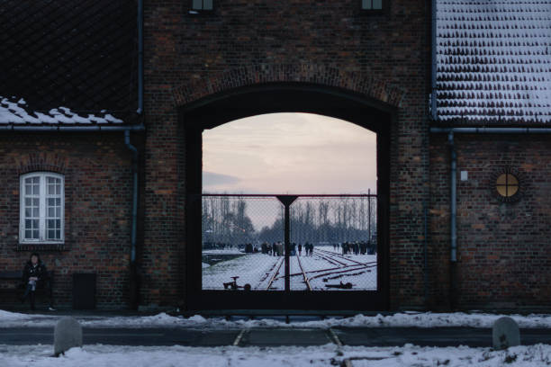 Auschwitz, Poland - November 30, 2023: The photo centers on Auschwitz’s entrance archway, revealing snow-covered rail tracks and a gathering of visitors paying tribute in the background. The setting sun casts a haunting glow, softening the stark lines of the gate and surrounding brick structures, creating a poignant, reflective mood.