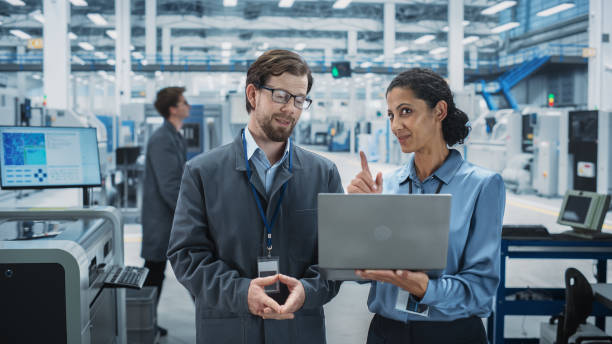 istock Caucasian Man and Hispanic Female Having a Conversation at Industrial Facility. Advanced Factory Employees Working Together, Using Laptop Computer for Supervising Work Process At Assembly Line. 2189597006