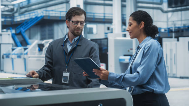 istock Male Technician and Hispanic Businesswoman Using Tablet Computer In Modern Electronics Factory. Caucasian Man Explaining Production Of Motherboards For AI-Powered Devices With Soldering Jet Printer. 2189597743