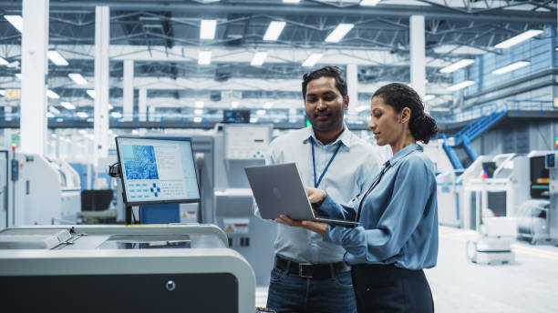 istock Indian Male Engineer And Hispanic Female Manager Using Laptop Computer And Talking At Electronics Factory. Man Using Soldering Jet Printer, Explaining Process Behind Production Of Circuit Boards 2189598481