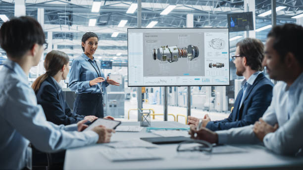 istock Electronics Factory Office Meeting: Hispanic Female Project Manager Presenting Innovative Engine On TV. Engineers And Technicians Listening, Taking Notes, Generating Ideas For the Production. 2189599048