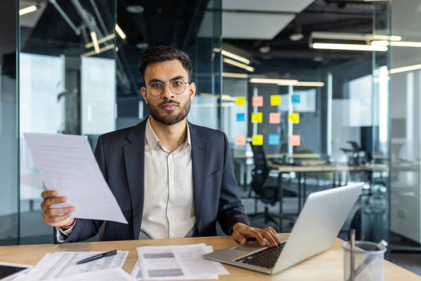 istock Confident businessman holding documents in modern office environment with laptop and papers on the desk, expressing focus and determination for corporate tasks and meetings 2189601256