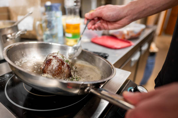 A chef bastes a seared steak in a shiny pan, enhancing its flavor with butter and herbs. The image captures the intricate process of gourmet cooking in a lively kitchen setting.
