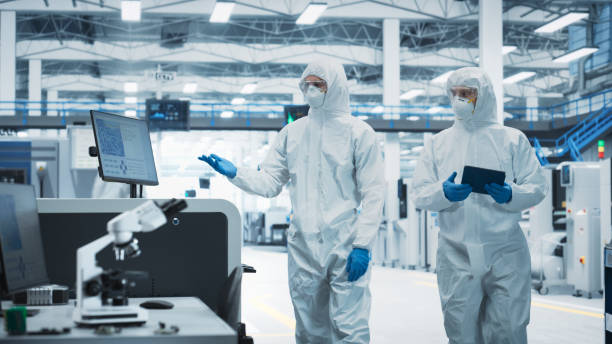 istock Two Scientists in Sterile Suits Walking In Electronics Factory Research Center. Technician With Tablet And Engineer Approaching Soldering Jet Printer And Starting Production Process Of Circuit Boards. 2189603585