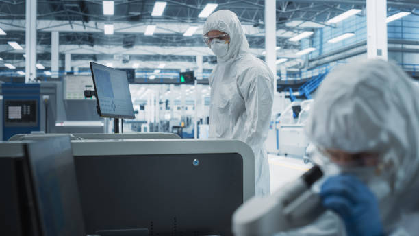 istock Two Scientists In Sterile Suites At Research Factory: Engineer Using Microscope And Desktop Computer To Inspect Microchip While His Colleague Using Soldering Jet Printer To Print Modern Motherboards. 2189604156