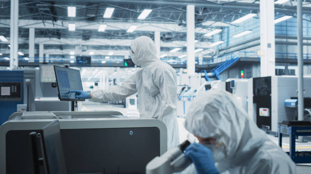istock Two Scientists In Sterile Suites At Research Factory: Engineer Using Microscope And Desktop Computer To Inspect Microchip While His Colleague Using Soldering Jet Printer To Print New Circuit Boards. 2189604186