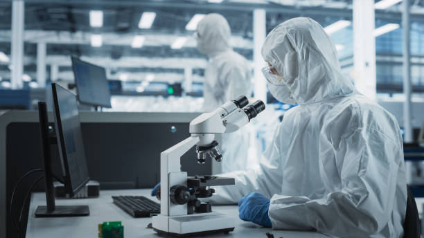 istock Two Scientists In Sterile Suites At Research Factory: Engineer Using Microscope And Desktop Computer To Inspect Microchip While His Colleague Using Soldering Jet Printer To Print Circuit Boards. 2189604222