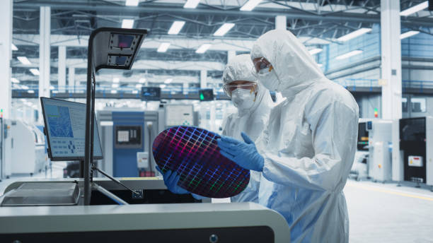 istock Semiconductor Production At Electronics Manufacturing Facility: Two Specialists In Sterile Suits Taking Out Silicon Wafer From Soldering Jet Printer And Inspecting. Engineers Making Computer Hardware 2189604306