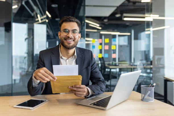 istock Confident businessman in modern office holding an envelope, smiling at the camera, with a laptop and tablet on the desk. Vibrant workspace with colorful notes in the background. 2189606331