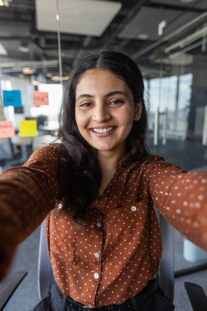 istock Young hispanic business woman smiling during a video call in modern office setting, using smartphone for professional communication and connectivity, dressed in casual polka-dot shirt 2189608371