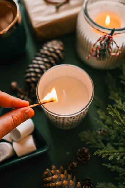 istock A hand lighting a festive candle with a matchstick, surrounded by pinecones, marshmallows, and evergreen branches, creating a warm and cozy Christmas atmosphere 2189609674