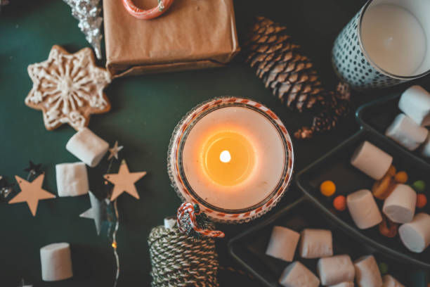 istock Cozy Christmas setup featuring a wrapped gift with a candy cane, a lit candle, tea, an open book, pinecones, marshmallows, and evergreen branches on a green background 2189609688