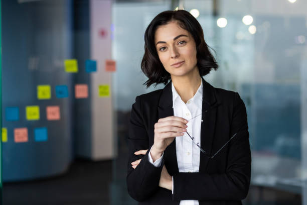 istock Confident businesswoman in modern office setting with sticky notes in background, wearing formal suit while holding glasses, ready for meeting, looking at camera with assurance 2189614482
