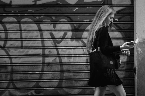 Florence, Italy - October 23, 2023: A young woman in contemporary attire walks along a graffiti-covered wall. Her focus on her phone contrasts with the artistic chaos of the urban background, a modern vibe amidst an old city.