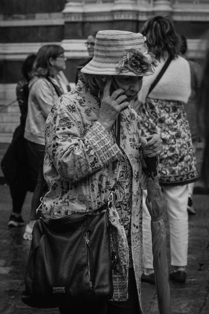 Florence, Italy - October 23, 2023: An elderly woman stands out with her patterned jacket and floral hat as she speaks on the phone. The vibrant individuality of her presence contrasts beautifully with Florence’s historic architecture in the background, showcasing the human connection within a city steeped in history.