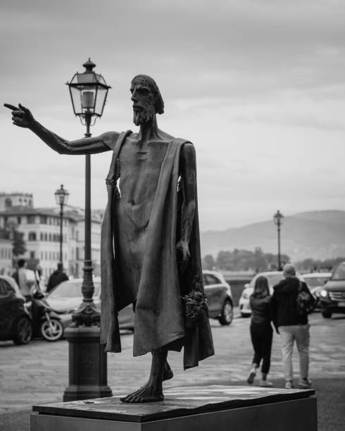 Florence, Italy - October 23, 2023: A majestic statue points toward the horizon, framed by elegant lampposts and a cloudy sky. The moody and atmospheric black and white photo highlights Florence’s historical charm and quiet reverence. People pass in the background, showcasing daily life amidst art.