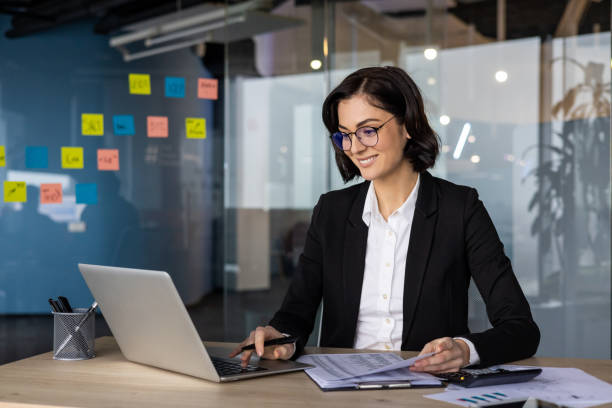 istock Confident businesswoman working on laptop in modern office with sticky notes displayed on glass wall, holding documents and calculator. Professional attire and focused expression indicate productivity. 2189617047