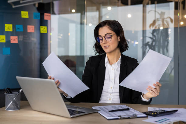 istock Young business woman analyzing documents at desk in modern office setting, wearing eyeglasses, working with laptop and reviewing important papers for strategic decision making 2189617368