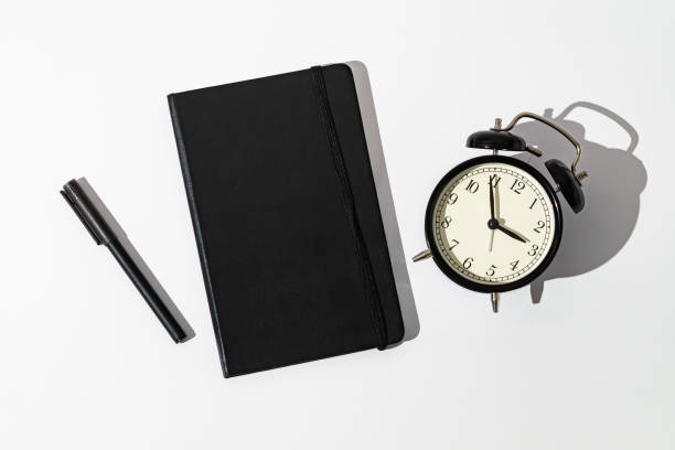 istock Black notebook, alarm clock and pen on white background. Planner for organization and goal priorities. Writing down tasks and schedule reminders on office desk. Minimal creative concept. Flat lay. 2189651323
