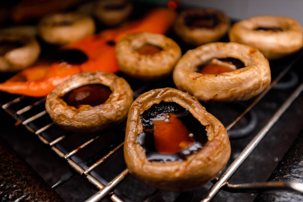 Mushrooms grilling on a electric grill, giving off a savory aroma. The heat from a electric grill enhances their flavor as they cook to perfection. Colorful peppers accompany them on the grill.