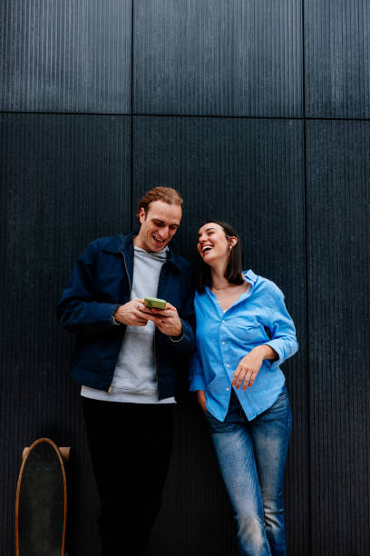 istock Young couple laughing while looking at smartphone against a wall 2189655492