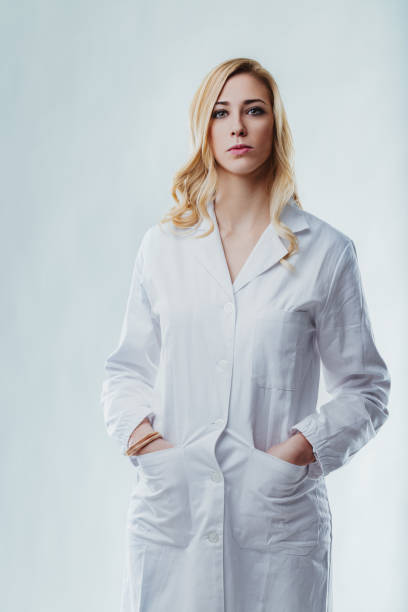 Portrait of a confident blonde female scientist wearing a lab coat and standing in her laboratory next to her equipment, looking at the camera with hands in her pockets