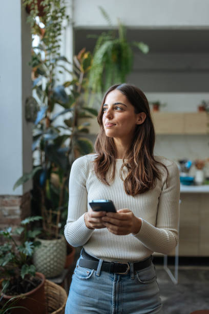 istock Young woman holding smartphone and looking away while standing next to window with plants 2189664138