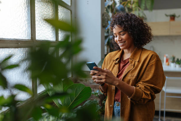 istock Young woman using mobile phone, standing next to window with plants 2189664177