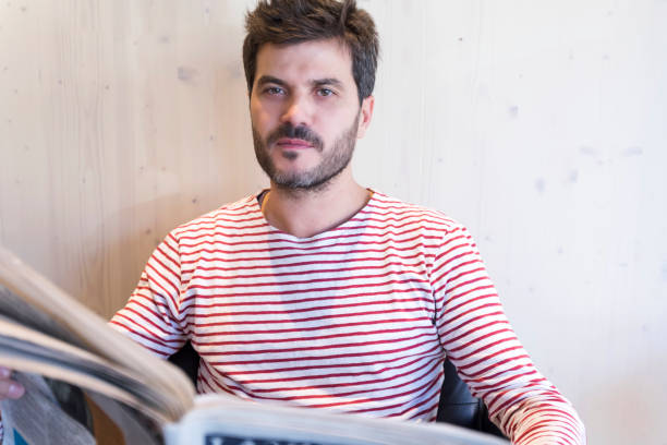 istock Man in a striped shirt reading a newspaper indoors. 2189723402