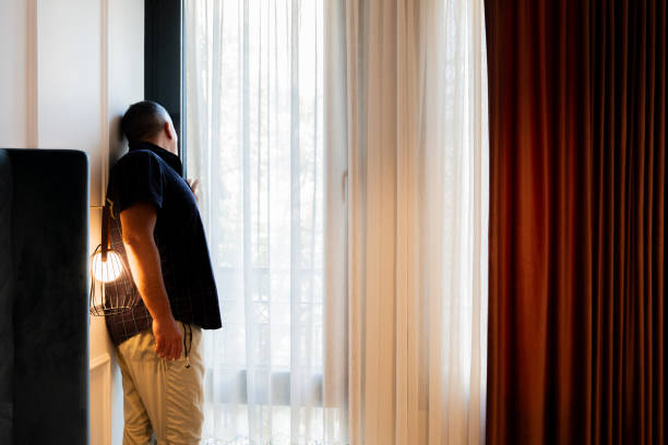 istock Man cautiously peering out of his hotel window to avoid being seen from outsideWomn 2189733297