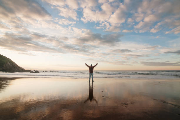 Rear view of a man with arms raised at a beach at sunset, and reflection on wet sand
