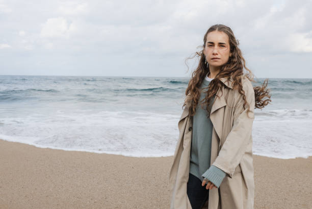 Young woman running on the beach to avoid waves. A young woman dressed in a trench coat runs along the beach shoreline avoiding the waves. walking beach women sadness stock pictures, royalty-free photos & images