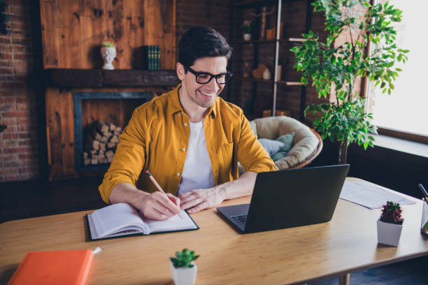 istock Young man enjoying leisure time at home, writing in notebook while smiling at laptop in stylish loft 2189753002