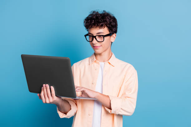 istock Young tech-savvy man with curly hair and glasses using laptop against blue background, showcasing modern lifestyle and stylish fashion 2189755023